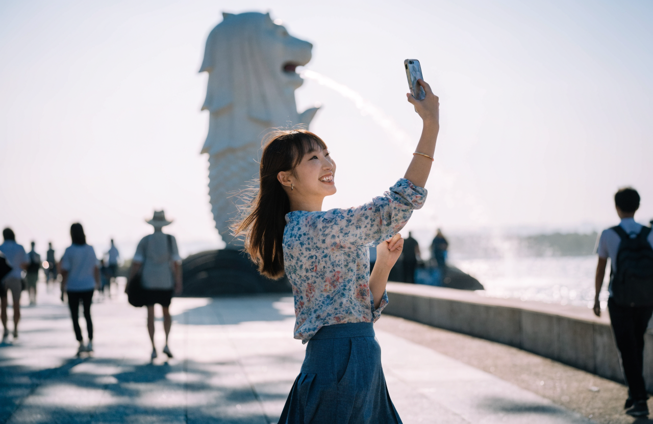 マーライオン公園の遊歩道に立つ笑顔の20代日本人女性が、マーライオン像を背景にスマートフォンで写真を撮ろうとしている場面